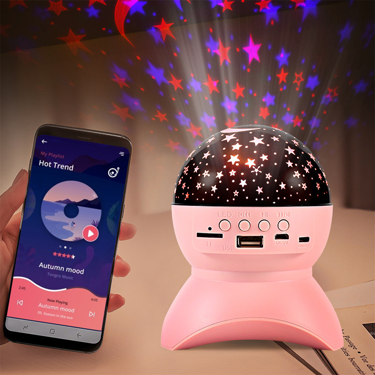 Rosa Sternprojektor, der Sterne projiziert. Telefon links spielt Musik. Weißer Hintergrund.