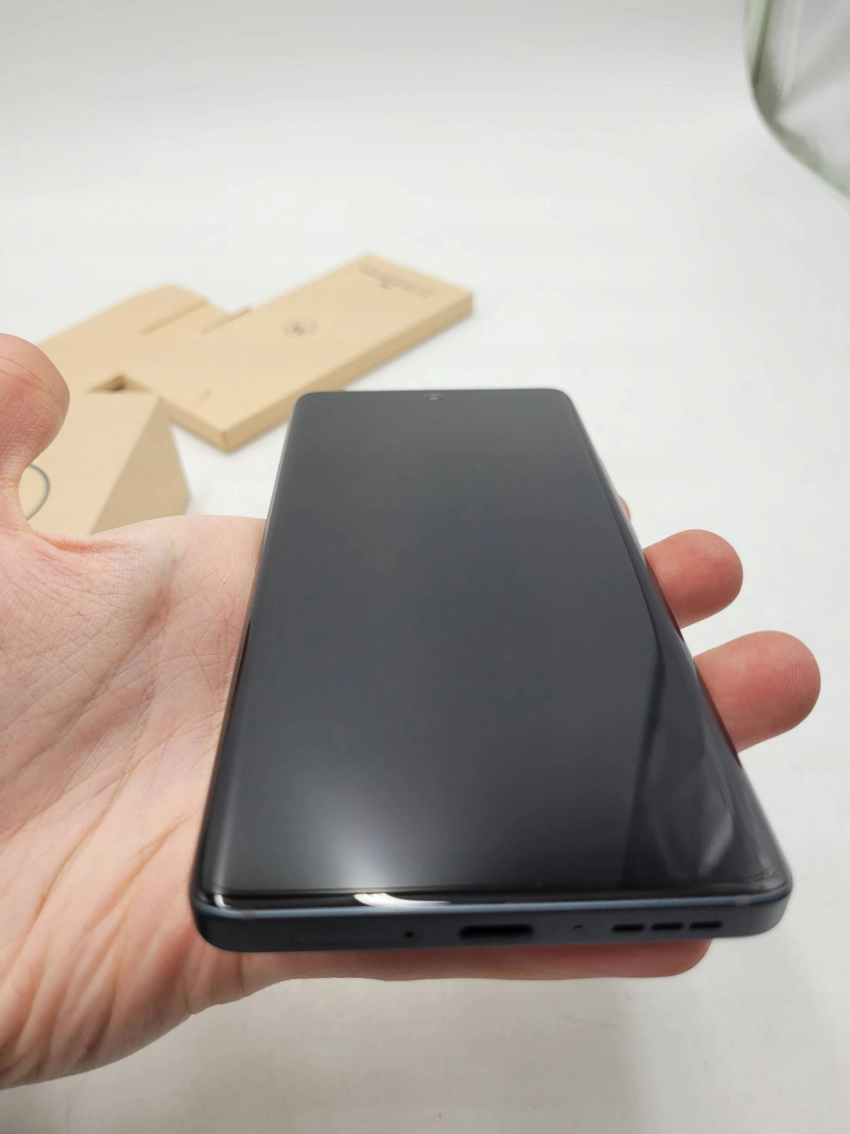 Eine Hand, die ein schwarzes Smartphone hält. Im Hintergrund sind einige Pappkartons.