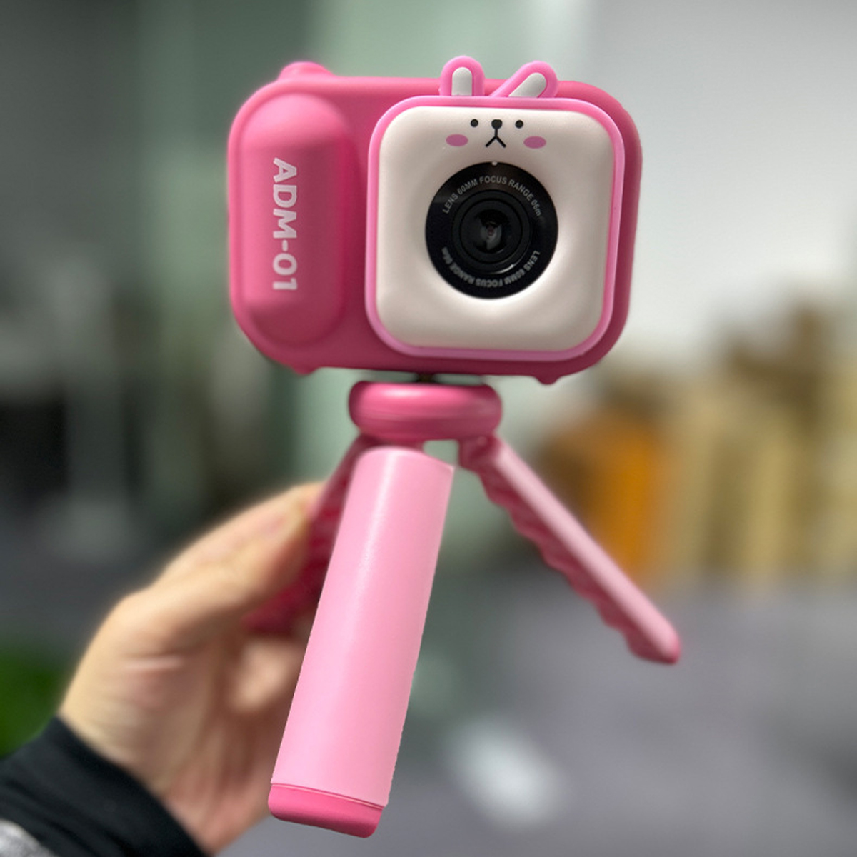 Różowy aparat z uszami królika. Aparat ma różowy uchwyt i jest trzymany przez rękę. Aparat ma obiektyw.