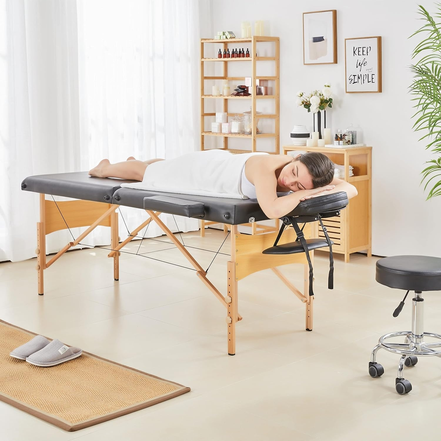 Frau auf Massagetisch, weißes Laken, neutraler Raum, Holzregal, Kunst.