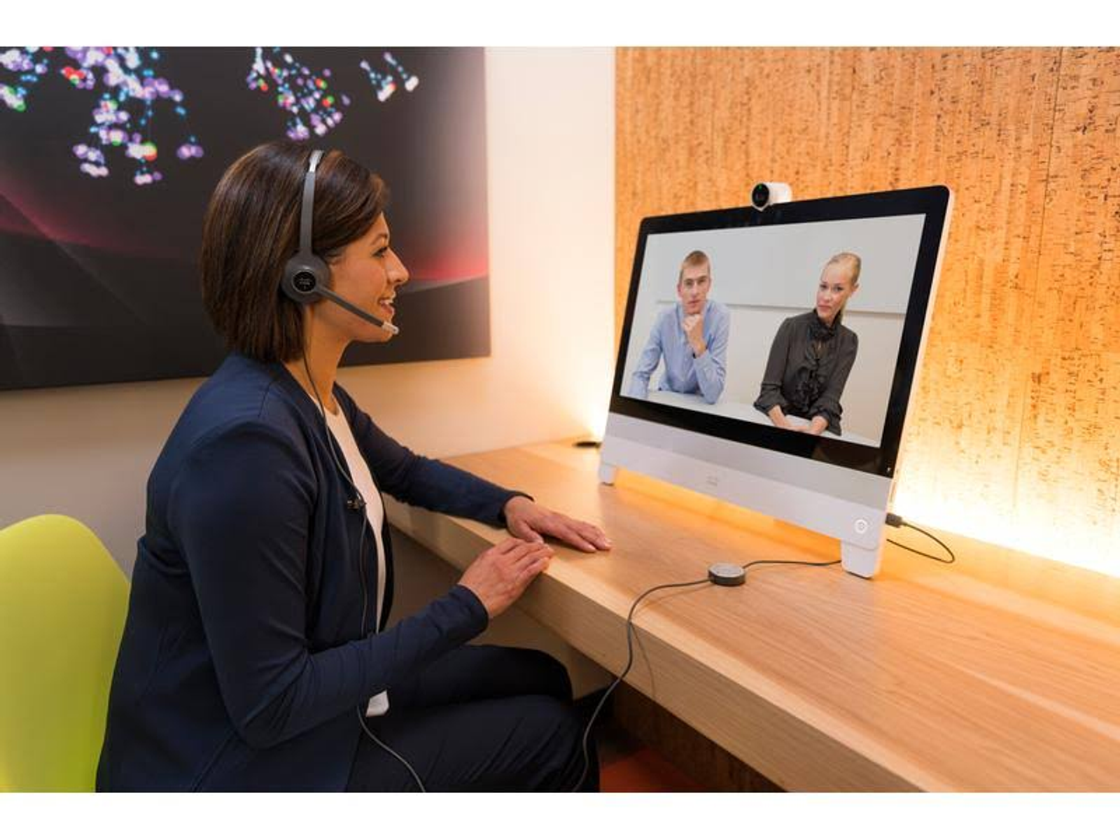 Kobieta podczas rozmowy wideo. Nosi zestaw słuchawkowy, siedzi przy biurku, na ekranie dwie osoby.