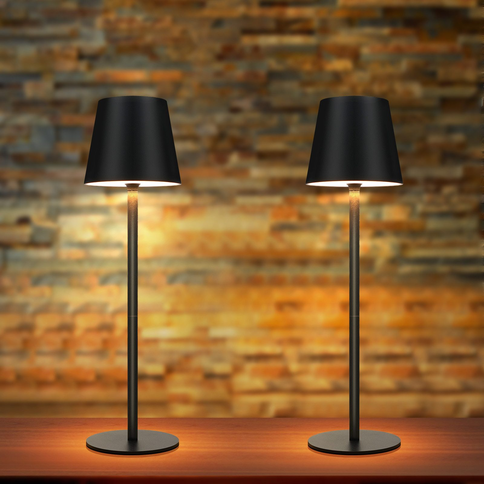 Dwie czarne lampy z czarnymi abażurami na drewnianym stole. Tłem jest ceglana ściana.