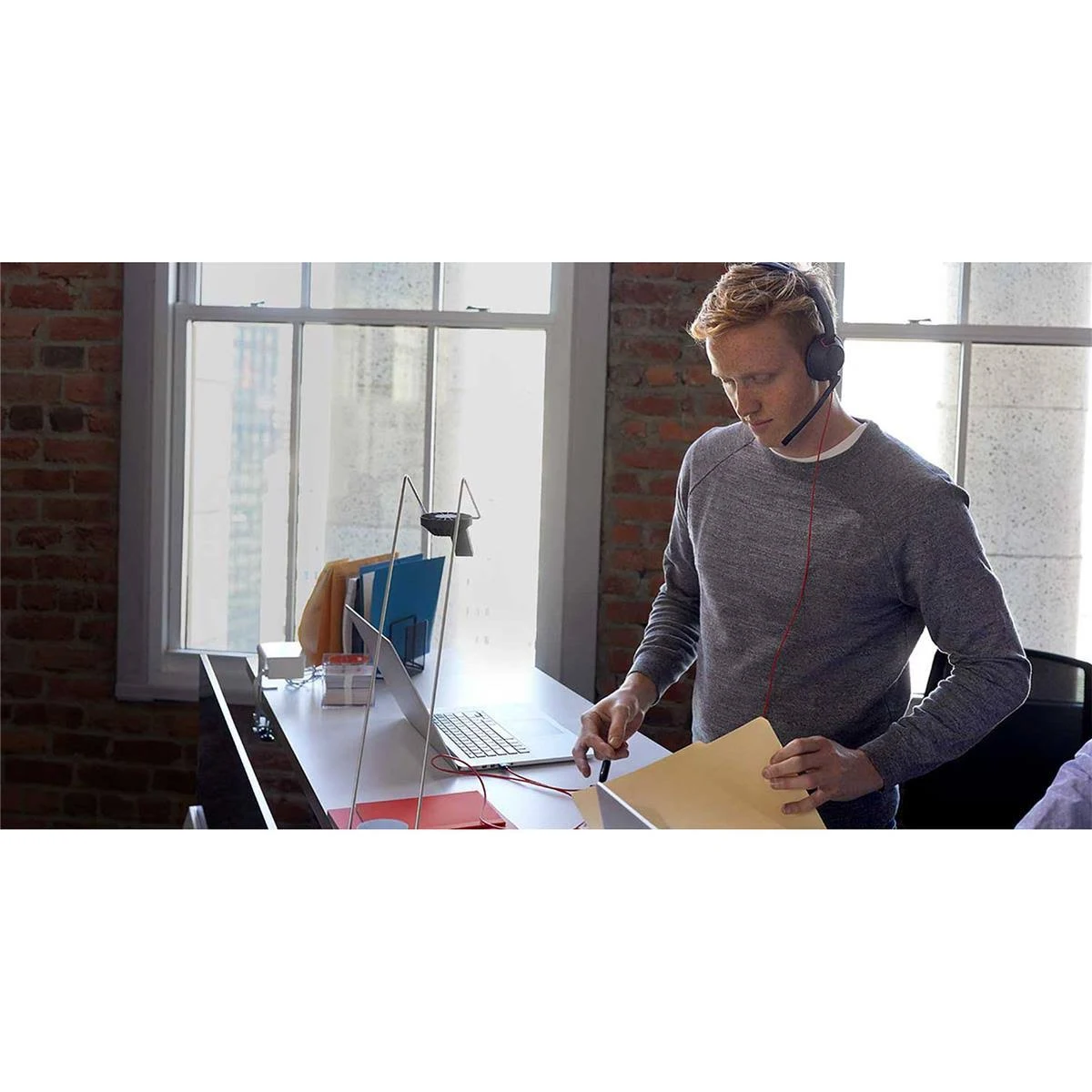 Mężczyzna w zestawie słuchawkowym, pracujący przy biurku z laptopem, papierami i oknem w tle.