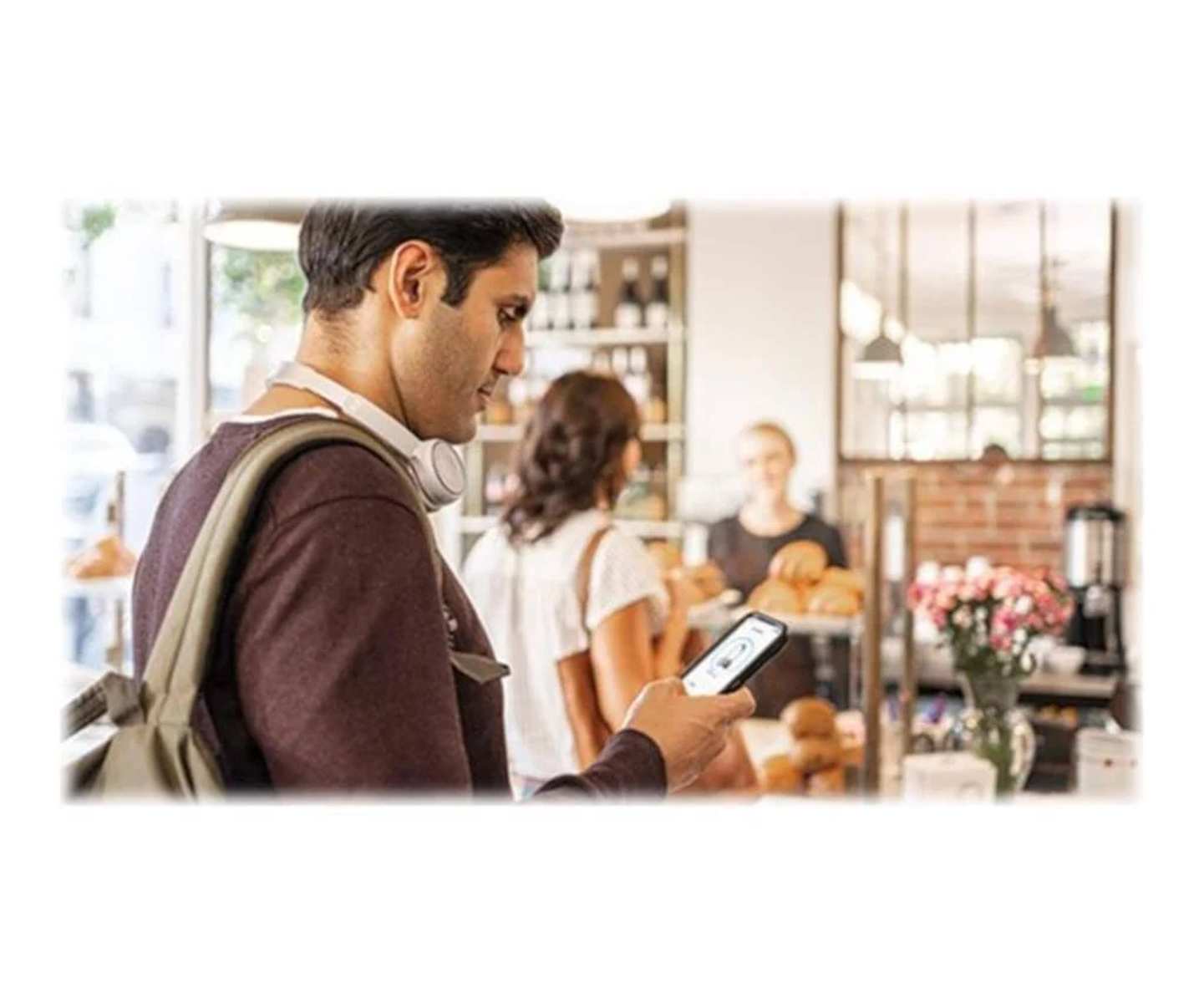 Mężczyzna w kawiarni patrzy na telefon. Kobieta i barista widoczni w tle.