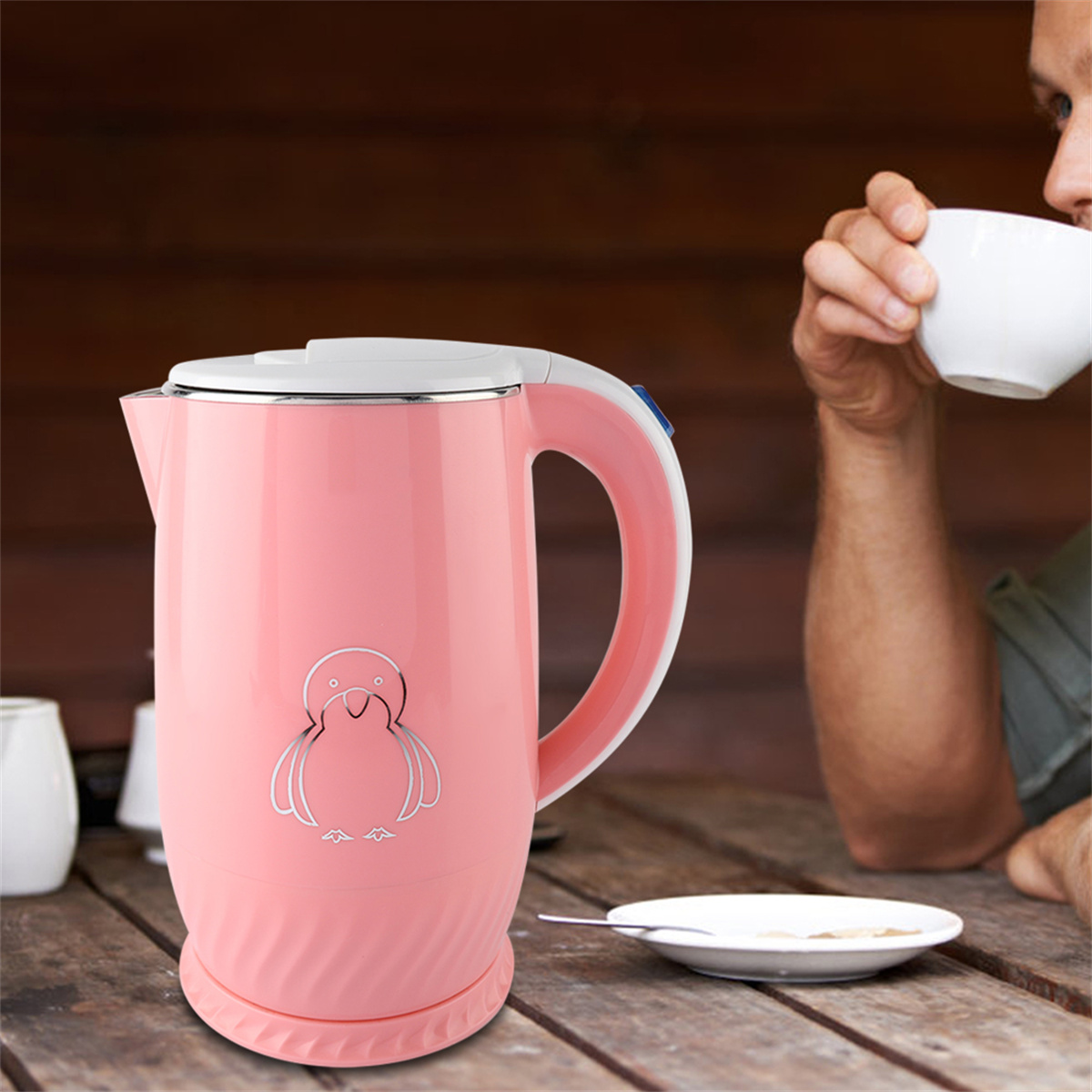 Rosa elektrischer Wasserkocher mit Pinguin-Design auf einem Tisch. Eine Person trinkt aus einer weißen Tasse.