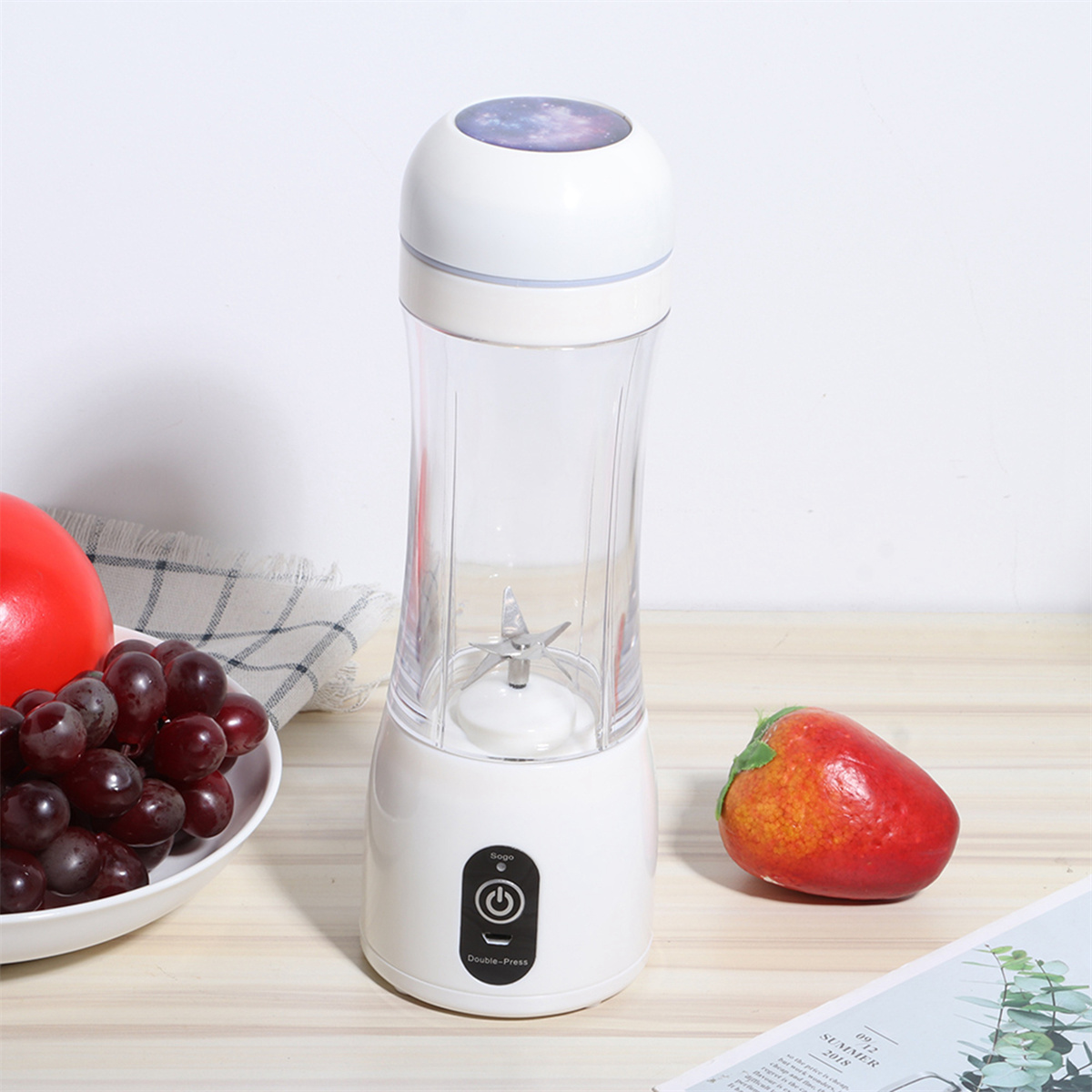 Ein weißer und klarer tragbarer Mixer. Eine Schüssel mit Trauben und eine rote Frucht sind links platziert.