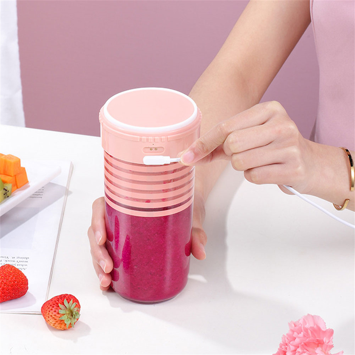 Frau steckt ein Ladekabel in einen rosa-roten Mixer. Erdbeeren und Früchte daneben.