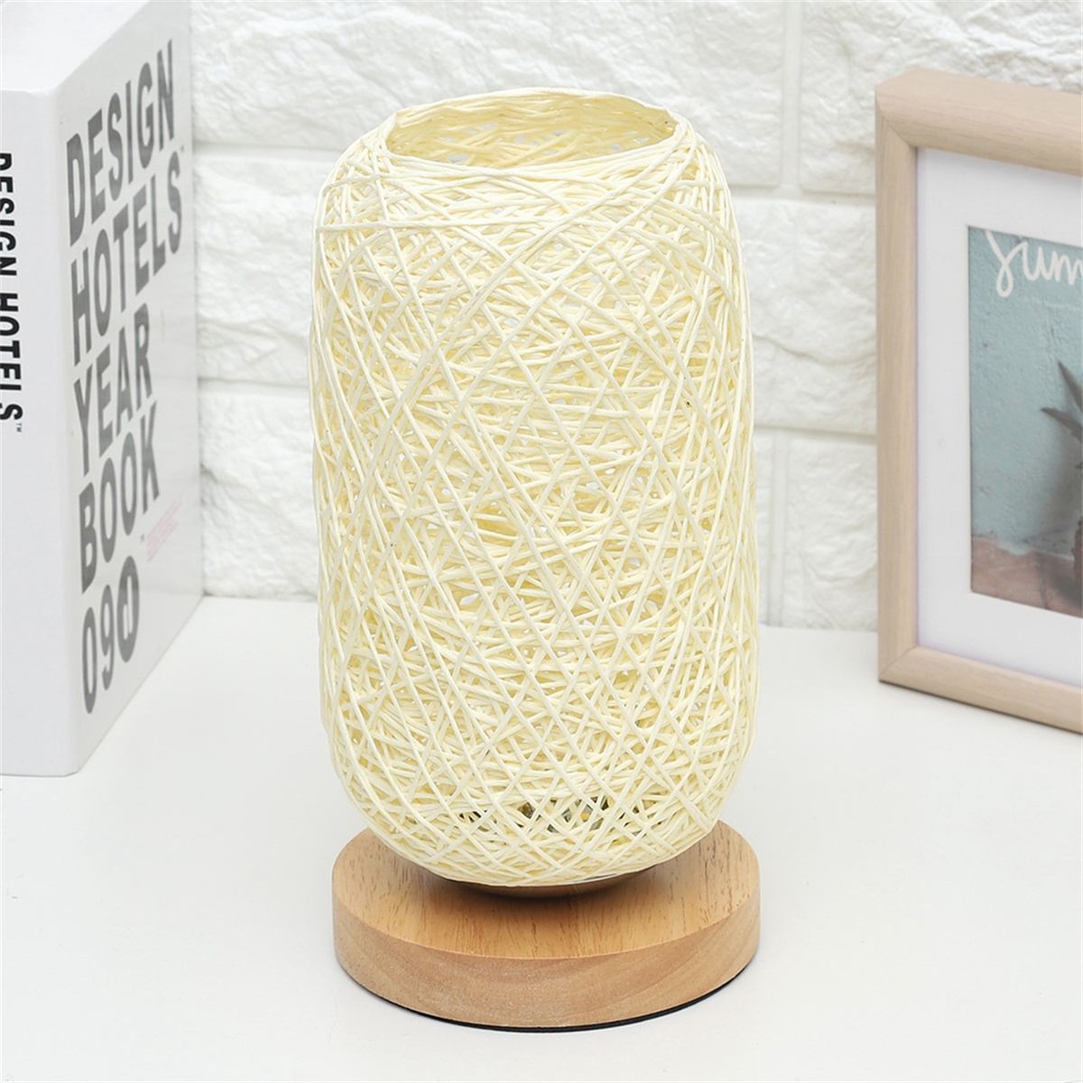 Cremefarbene gewebte Lampe auf Holzsockel, dahinter ein Designbuch und ein gerahmtes Bild.