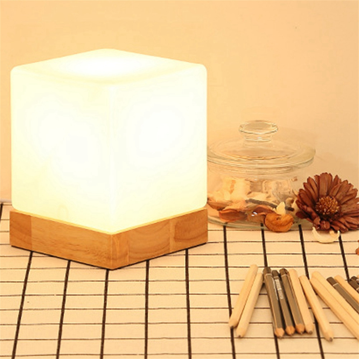 Beleuchtete würfelförmige Lampe auf Holzsockel. Ein Glas und Bleistifte stehen auf dem Tisch.