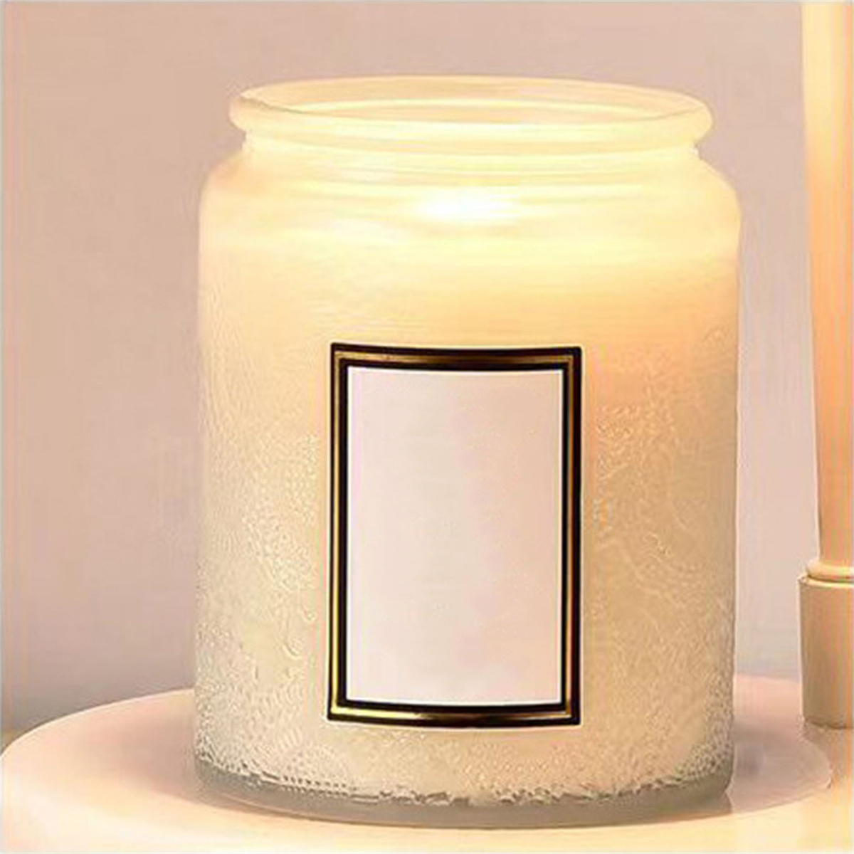 Angezündete Kerze in einem Glas. Sie hat weißes Wachs und ein Etikett. Das Licht ist gelblich.