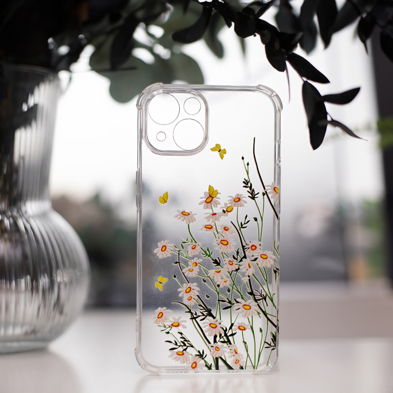 Transparente Handyhülle mit weißen Gänseblümchen und gelben Schmetterlingen. Die Hülle liegt auf einem Schreibtisch vor einem Fenster.