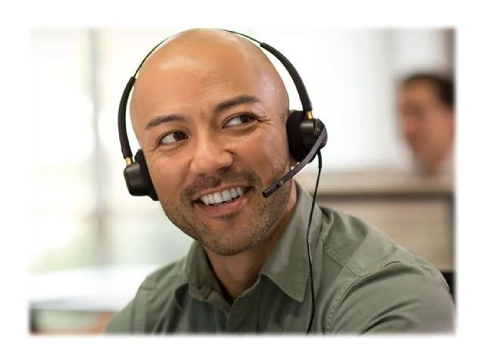 Uomo calvo con cuffie, sorridente, con uno sfondo sfocato di un ambiente ufficio.