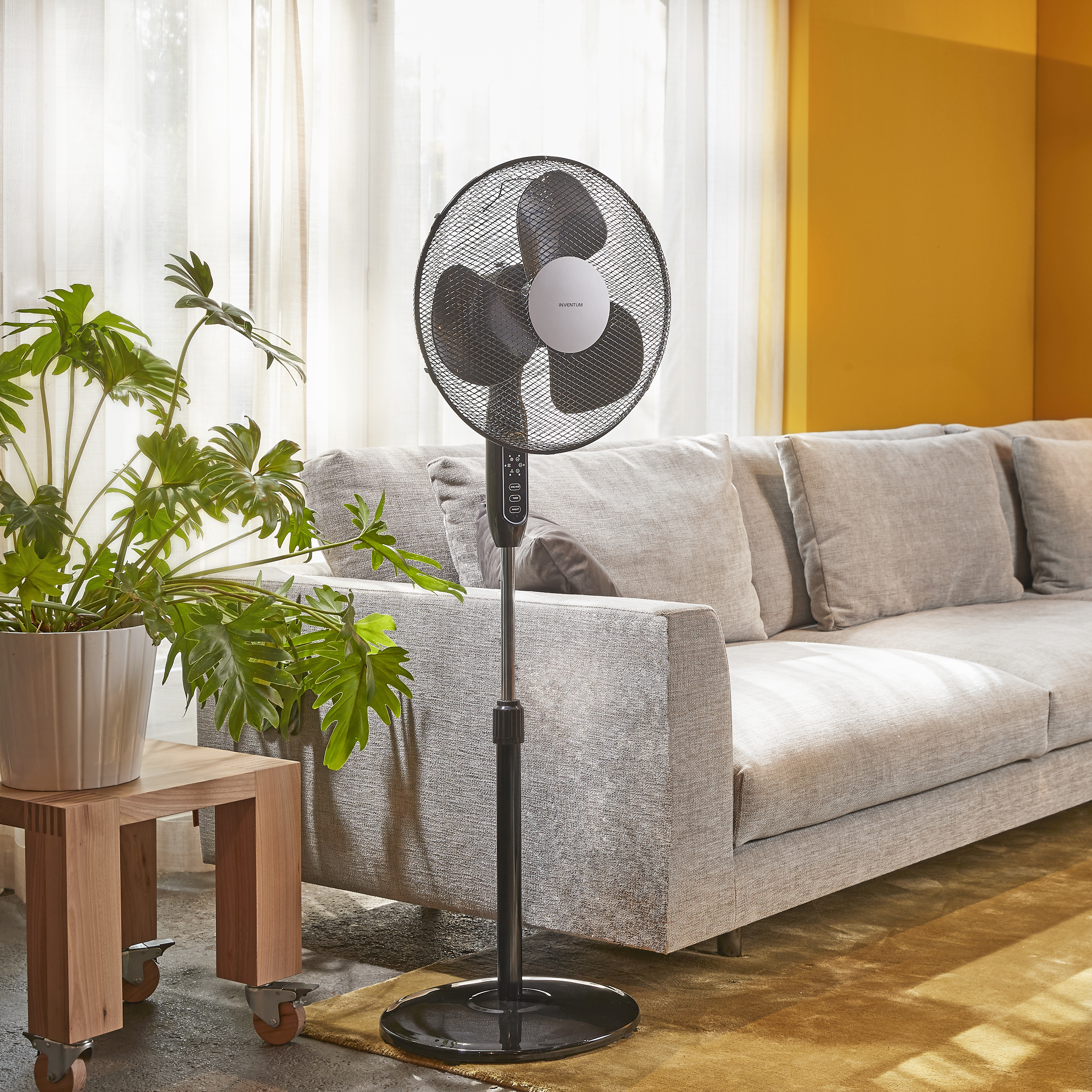 Un ventilateur noir et argenté sur pied, près d'un canapé et d'un mur jaune.