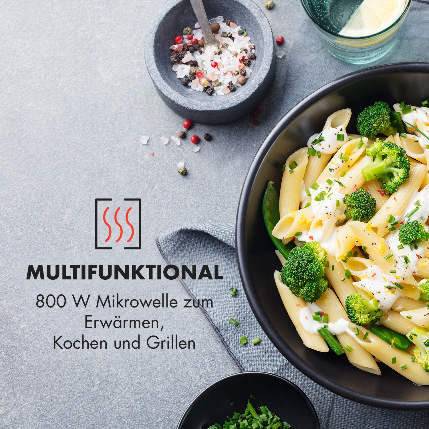 Eine Schüssel Pasta mit Brokkoli und Sahnesauce, neben einer Schüssel mit Gewürzen und Kräutern. Graue Oberfläche.