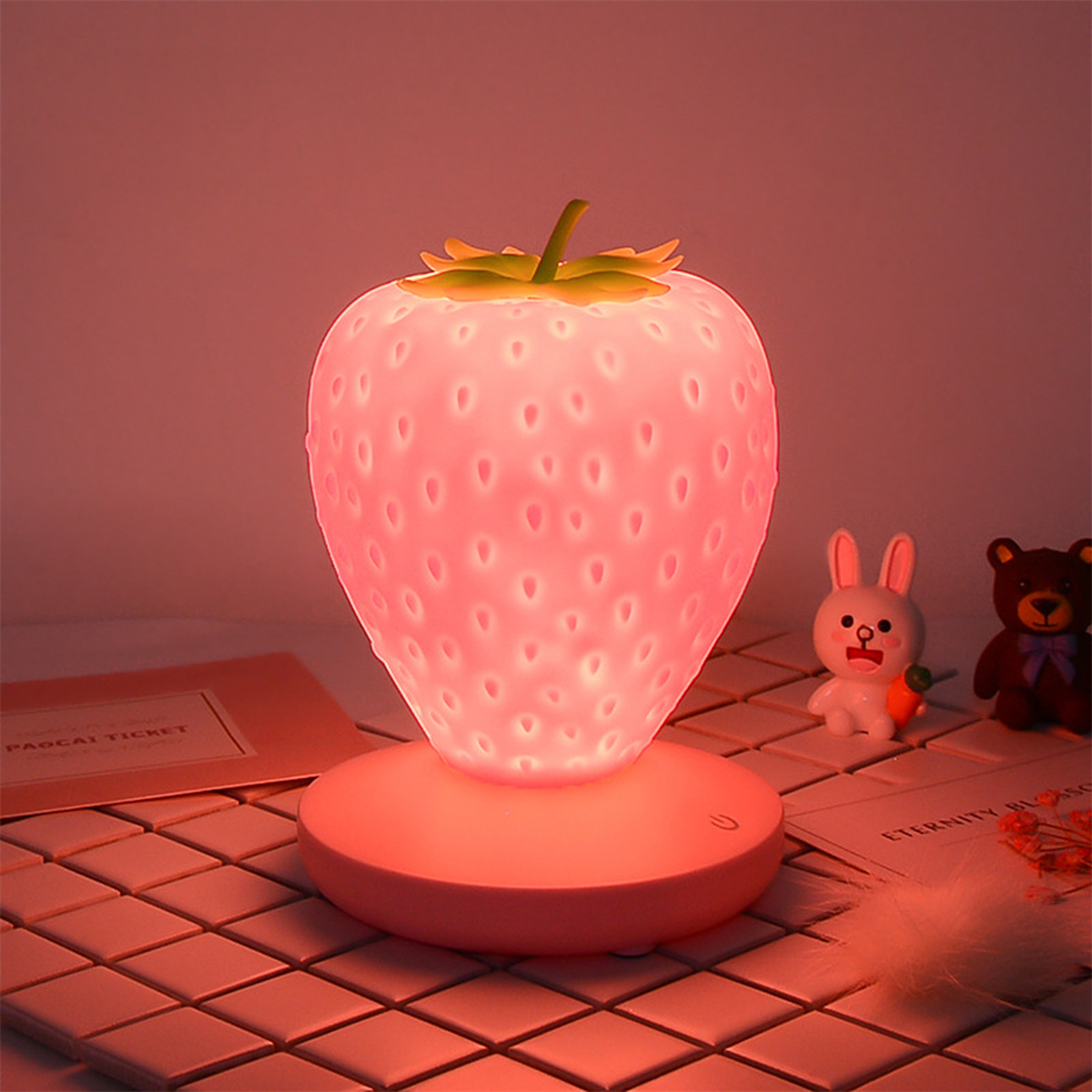 Erdbeerförmige Lampe, die rosa Licht aussendet, auf rosa Basis, mit Spielzeug im Hintergrund.