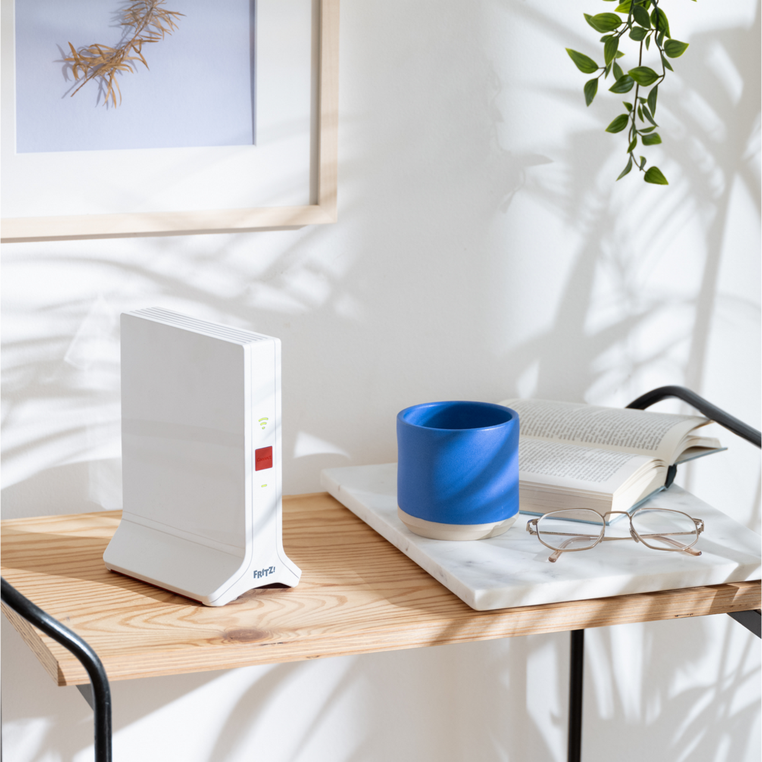 Biały router z czerwonym przyciskiem, niebieski kubek, książka i okulary na drewnianej półce.