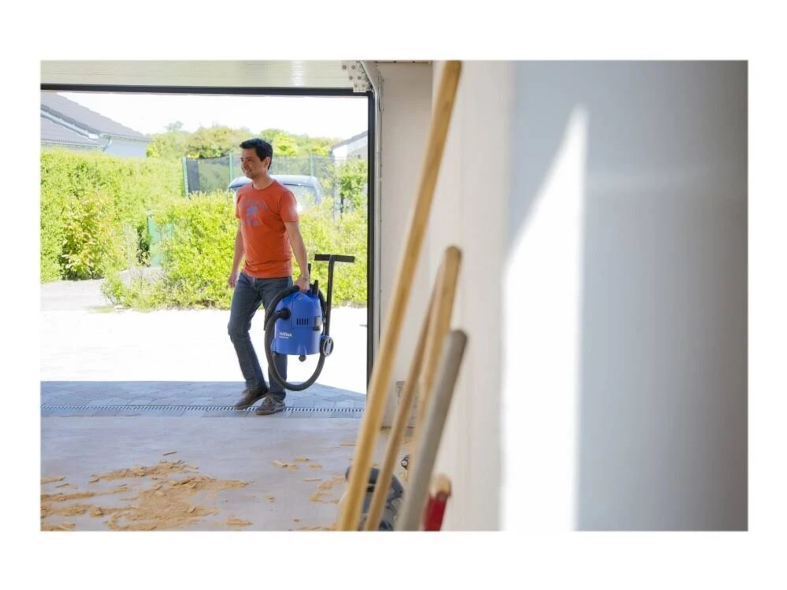 Mężczyzna w garażu niosący odkurzacz. Narzędzia widoczne w tle.