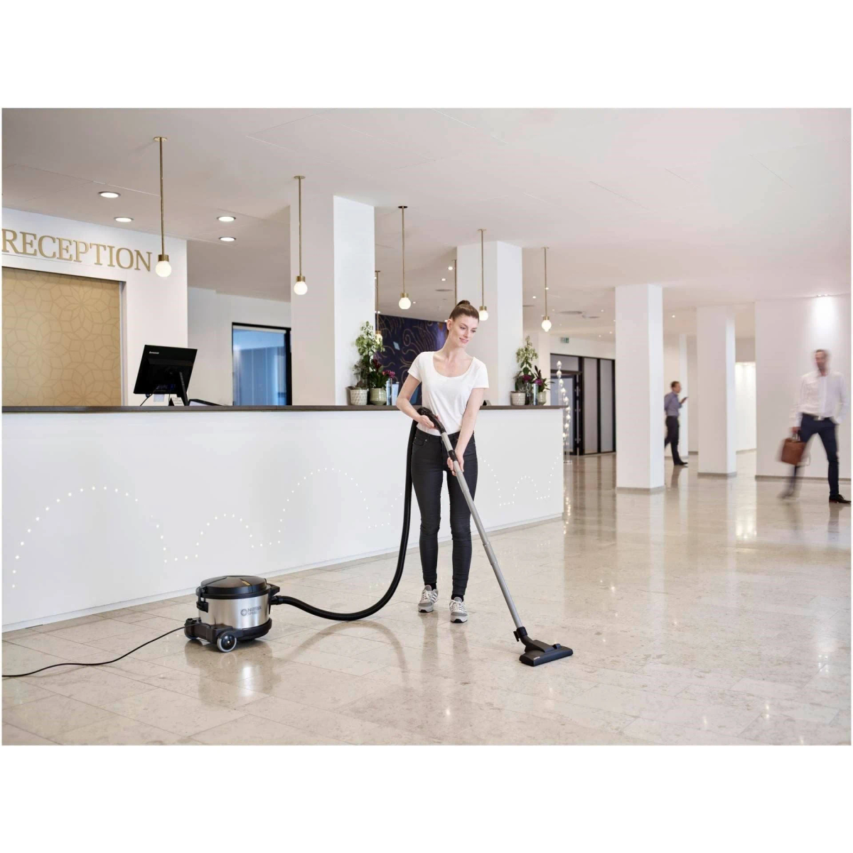 Mujer aspirando un vestíbulo de hotel. Recepción y otras personas en el fondo.