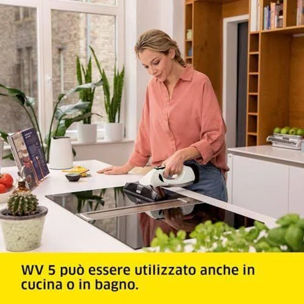 Una donna usa un aspirapolvere portatile su un piano cottura. Sfondo cucina con piante ed elettrodomestici.