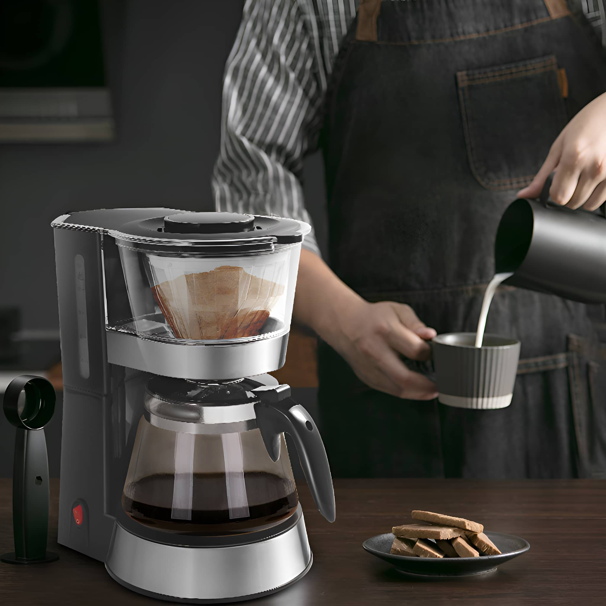Eine Person gießt Milch aus einem schwarzen Krug in eine Tasse neben einer Kaffeemaschine und einem Teller mit Keksen.