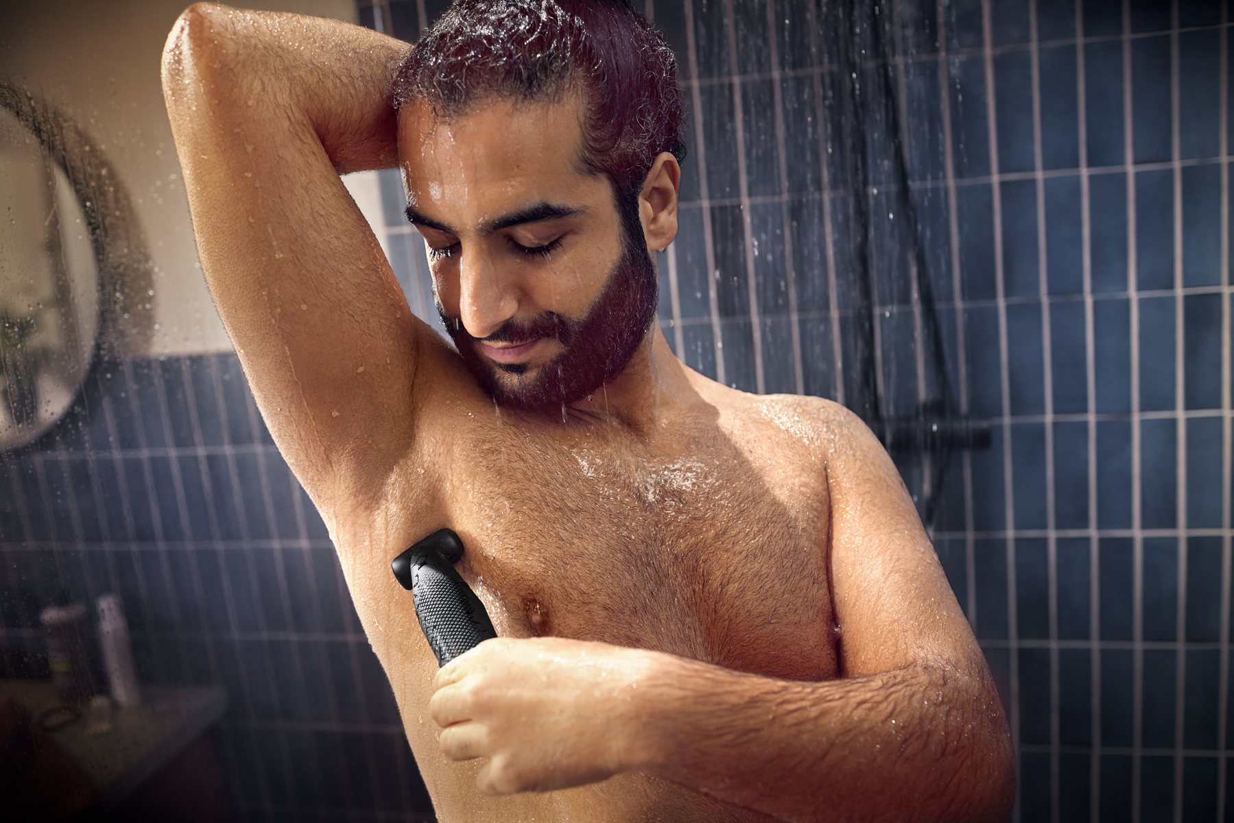 Man shaving his armpit with a trimmer in the shower, with a tiled wall background.