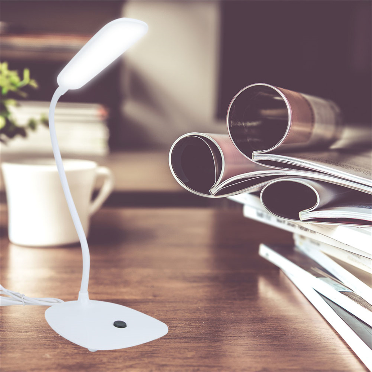 Eine weiße Schreibtischlampe mit gebogenem Hals, beleuchtet. Ein Stapel Zeitschriften und eine Kaffeetasse sind im Hintergrund.