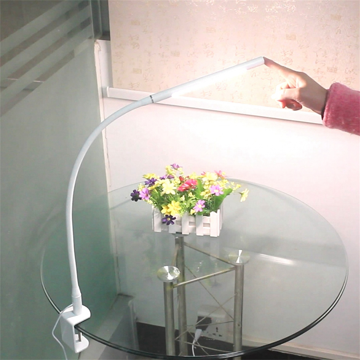 Eine weiße, gebogene Schreibtischlampe mit Klemme, die auf eine Hand zeigt. Darunter steht ein kleiner Blumenstrauß.