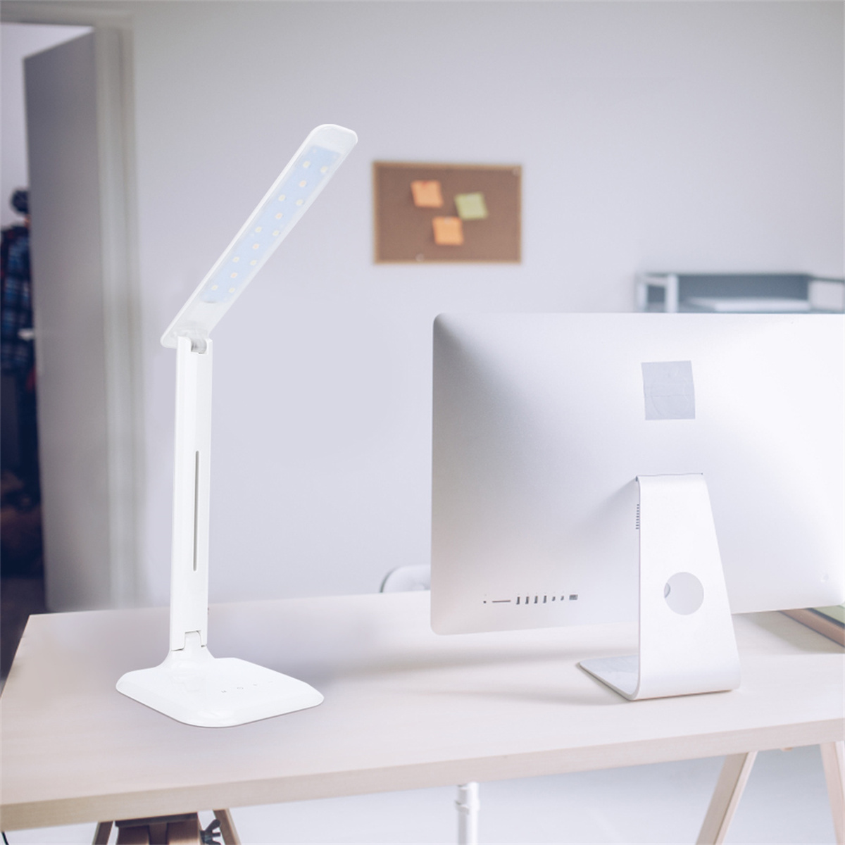 Weiße Schreibtischlampe auf einem Schreibtisch mit Computermonitor. Eine Pinnwand ist im Hintergrund.