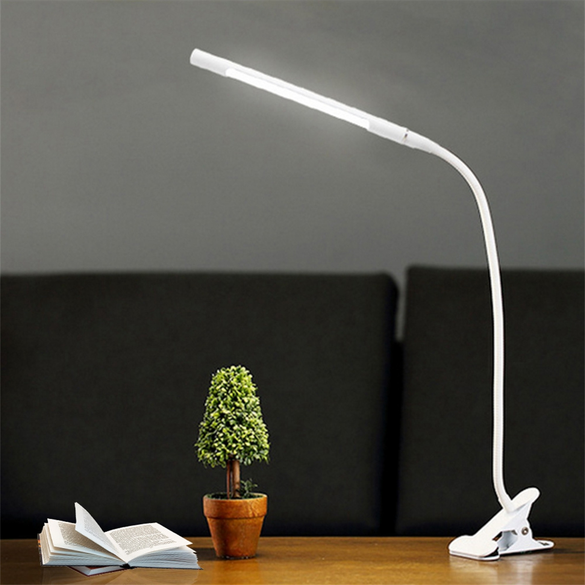 Eine weiße Schreibtischlampe mit flexiblem Arm, einer Pflanze und einem offenen Buch.
