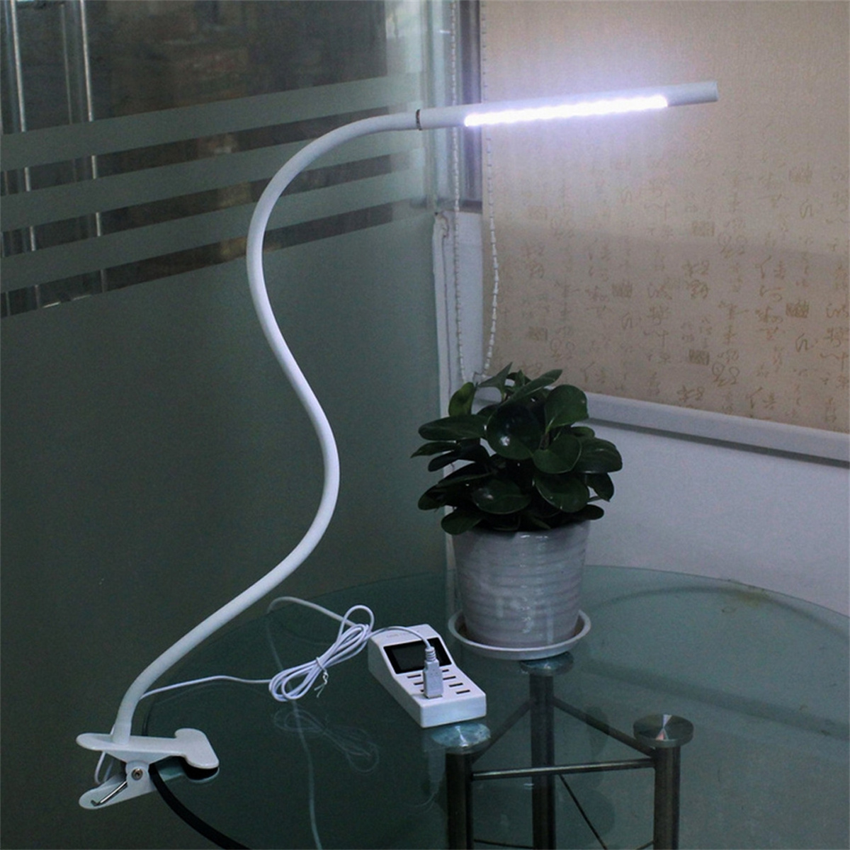 Weiße Schreibtischlampe und Pflanze. Die Lampe ist weiß, mit einem flexiblen Arm. Eine Pflanze rechts.