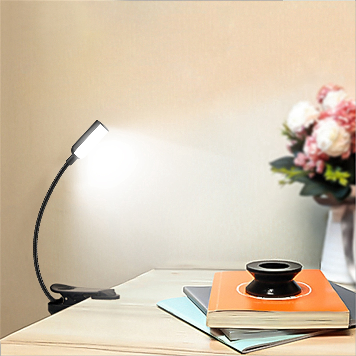 Eine schwarze Leselampe, die an einem Schreibtisch befestigt ist und Licht auf einen Stapel Bücher und eine Blumenvase wirft.