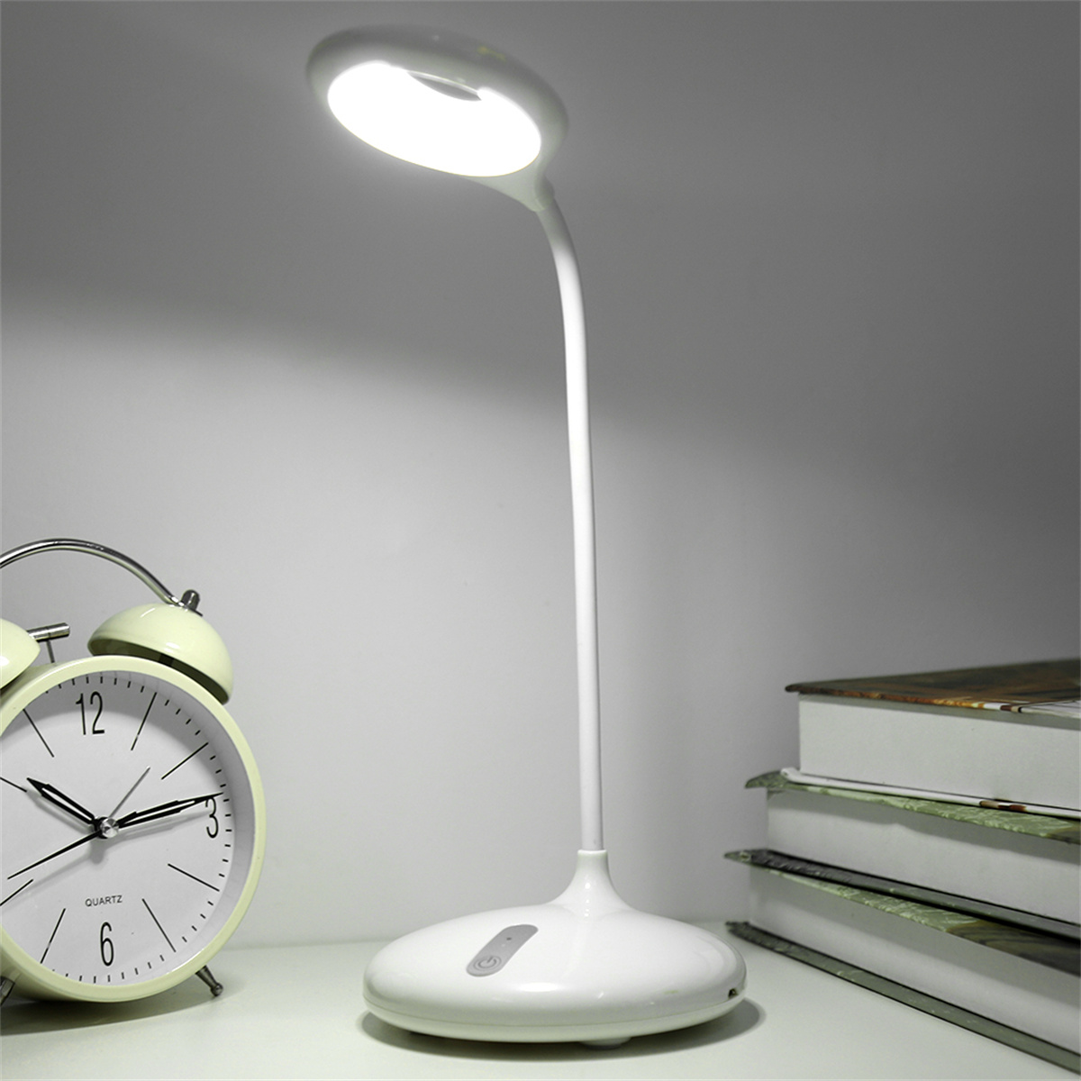Weiße Schreibtischlampe mit rundem Sockel und hellem Licht, Uhr und Bücher sichtbar.