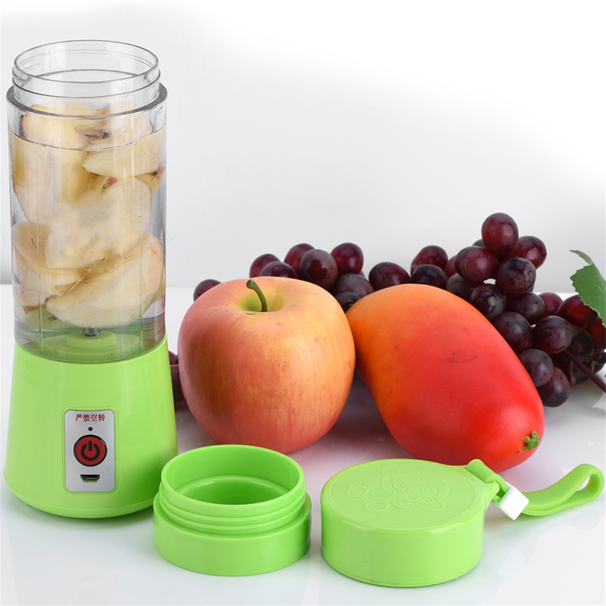 Tragbarer grüner Mixer mit Äpfeln und Mango darin. Trauben und eine weitere Mango liegen daneben.