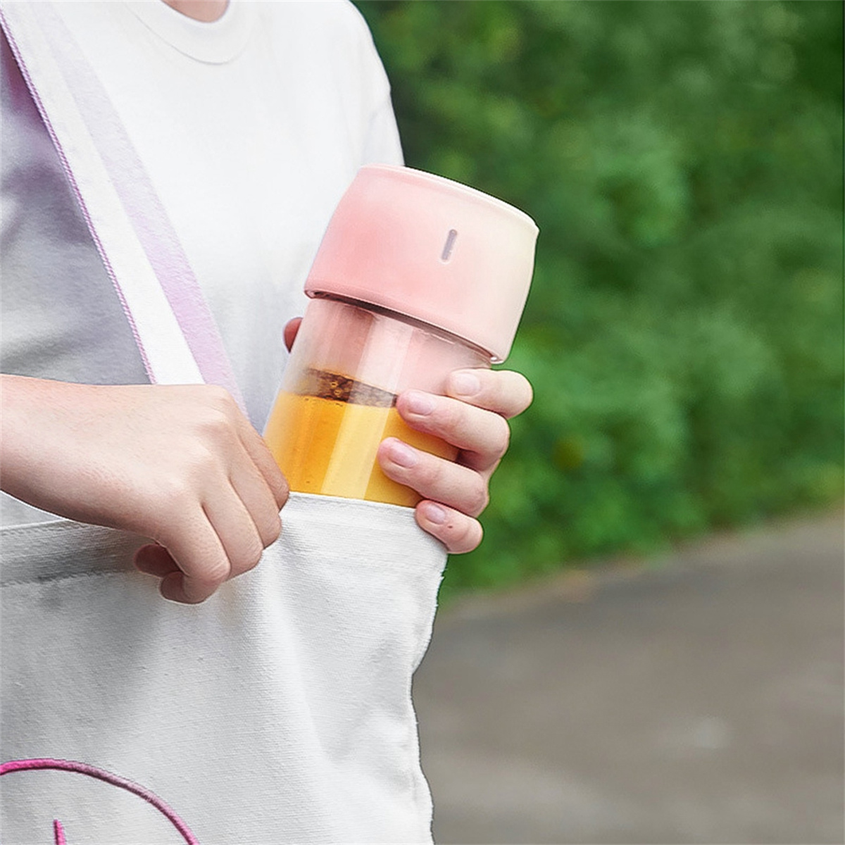 Eine Person hält einen rosa-klaren Mixer mit orangener Flüssigkeit, in einer weißen Tasche.