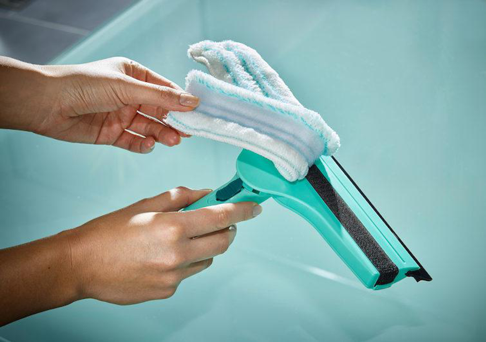 Dłonie osoby trzymającej zielono-białą myjkę do okien. Myjka na niebieskiej powierzchni.
