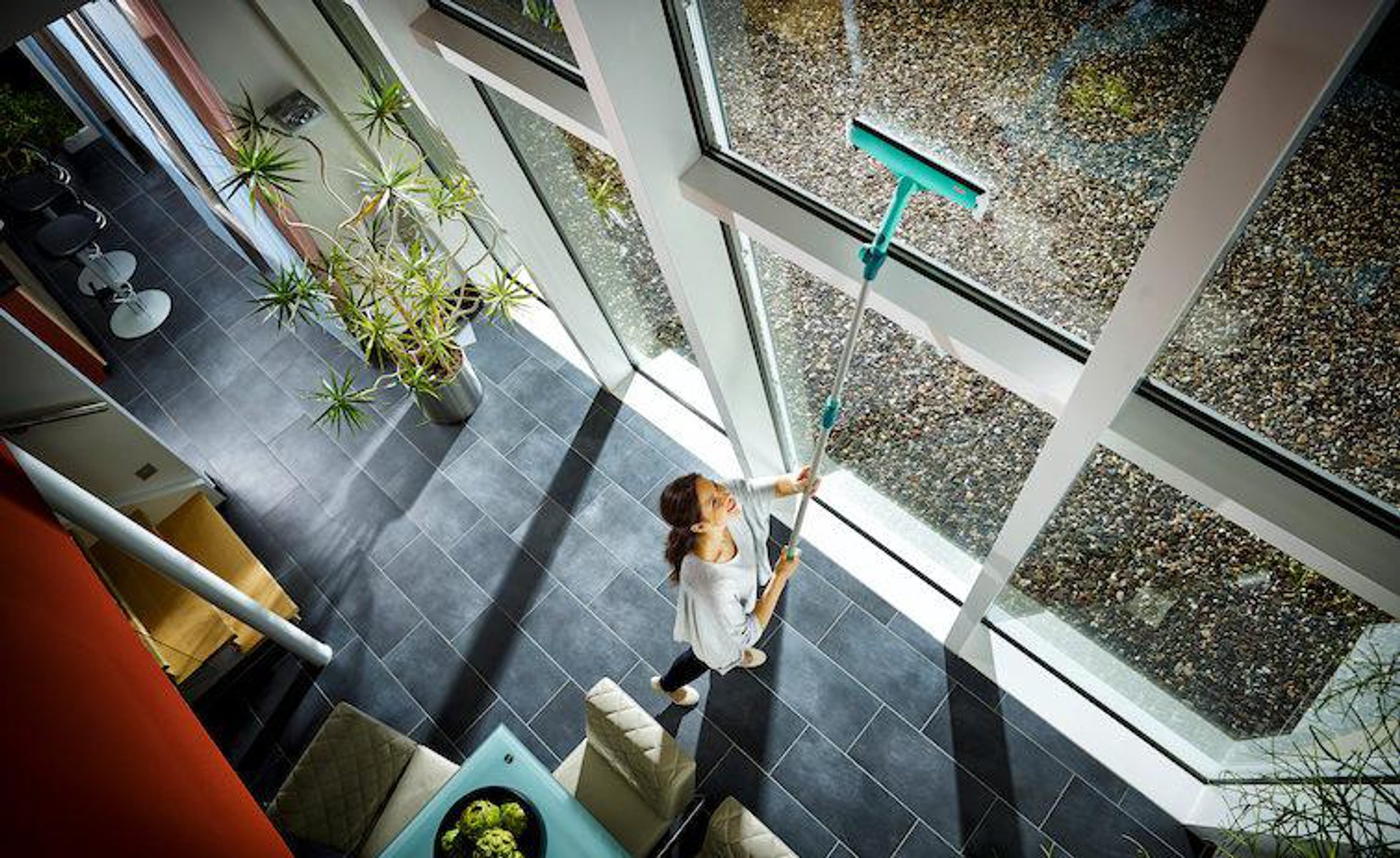 Kobieta myjąca duże okno za pomocą ściągaczki z długim uchwytem w dobrze oświetlonym, nowoczesnym pomieszczeniu.