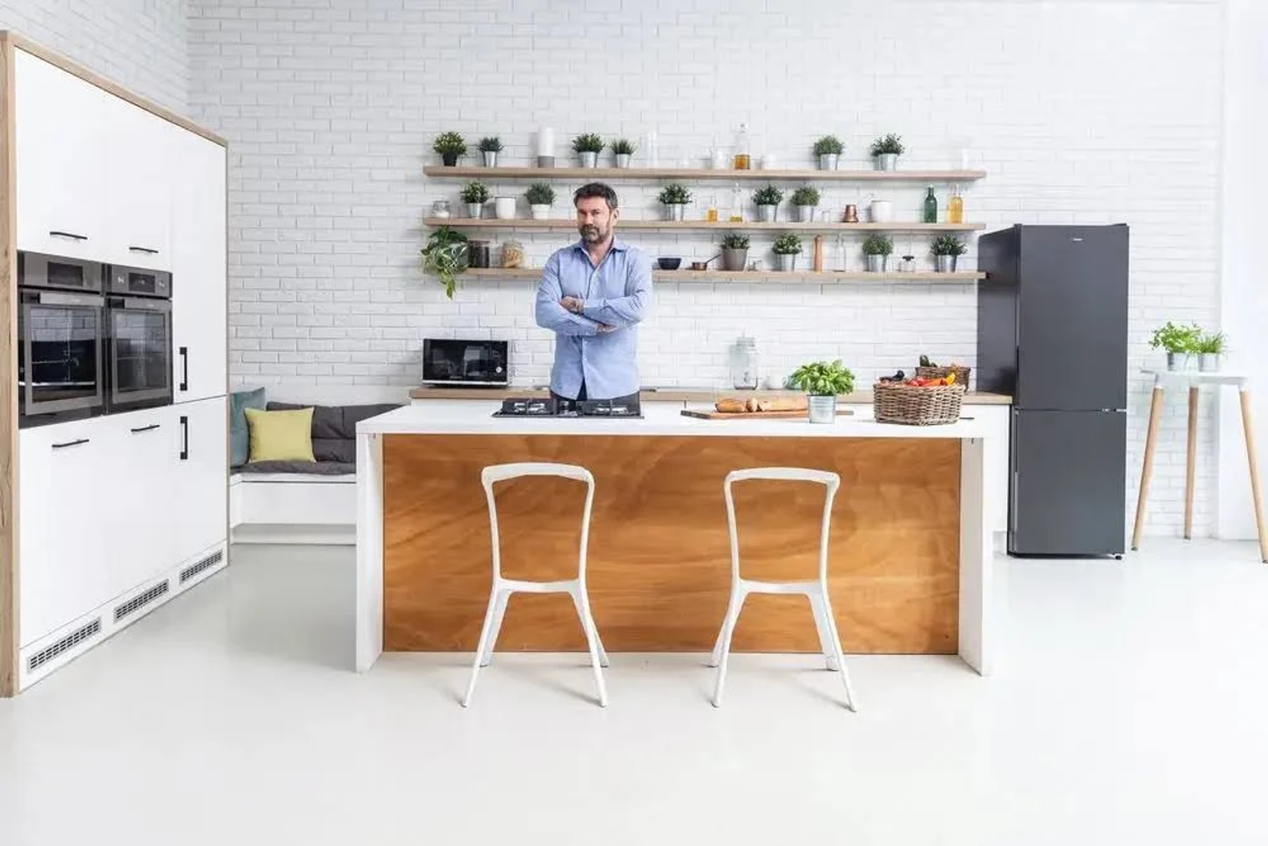Uomo in una cucina con mobili bianchi, parete in mattoni e un'isola in legno. Piante, cibo sono visibili.