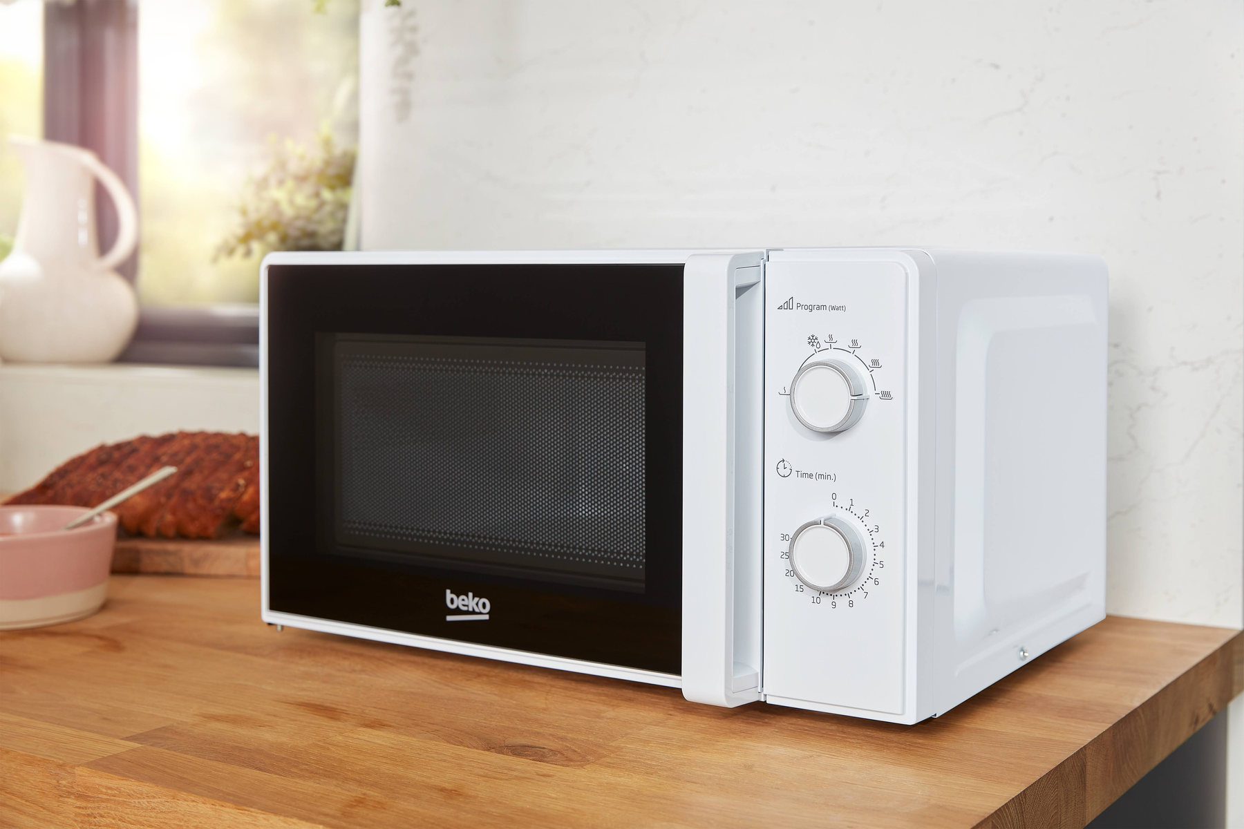 Biała kuchenka mikrofalowa Beko na drewnianym blacie. Sterowanie po prawej. Chleb i dzbanek w tle.