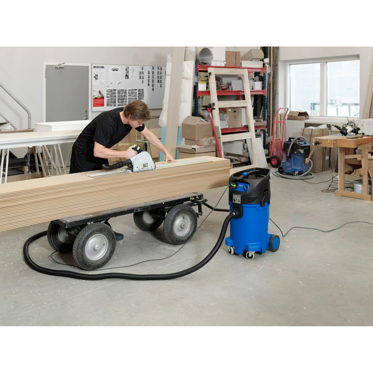Ein Mann schneidet Holz mit einer Säge. Es gibt einen Wagen mit Holz und einen blauen Staubsauger.