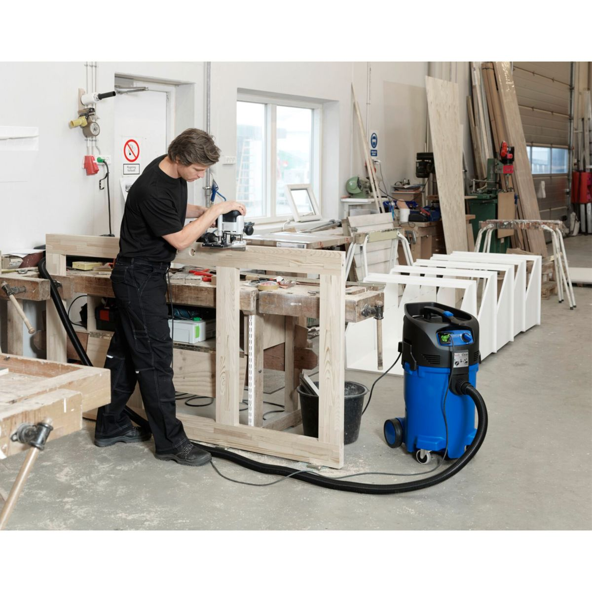 Ein Mann benutzt einen Router an einem Holzrahmen. Staubsauger und Werkzeuge befinden sich in der Werkstatt.