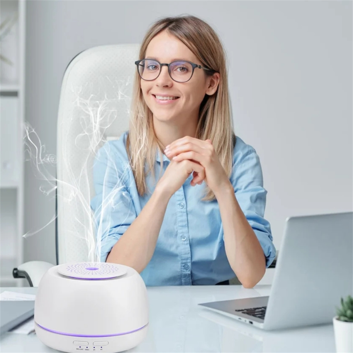 Eine Frau mit Brille lächelt vor einem weißen Diffusor, der Dampf abgibt, an einem Schreibtisch sitzend.