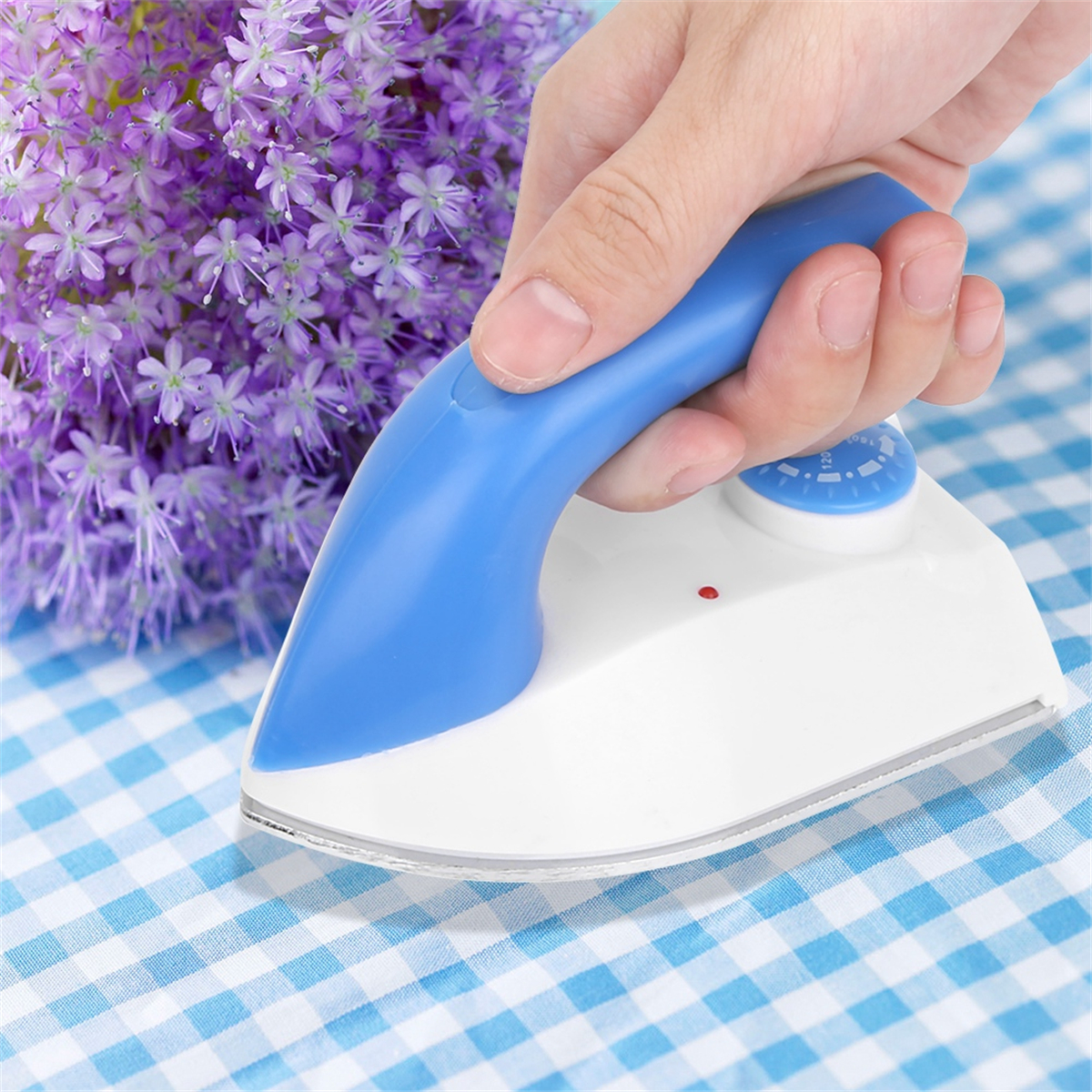 Ein kleines weißes Bügeleisen mit blauem Griff auf karierter Tischdecke, in der Nähe eines lila Blumenstraußes.