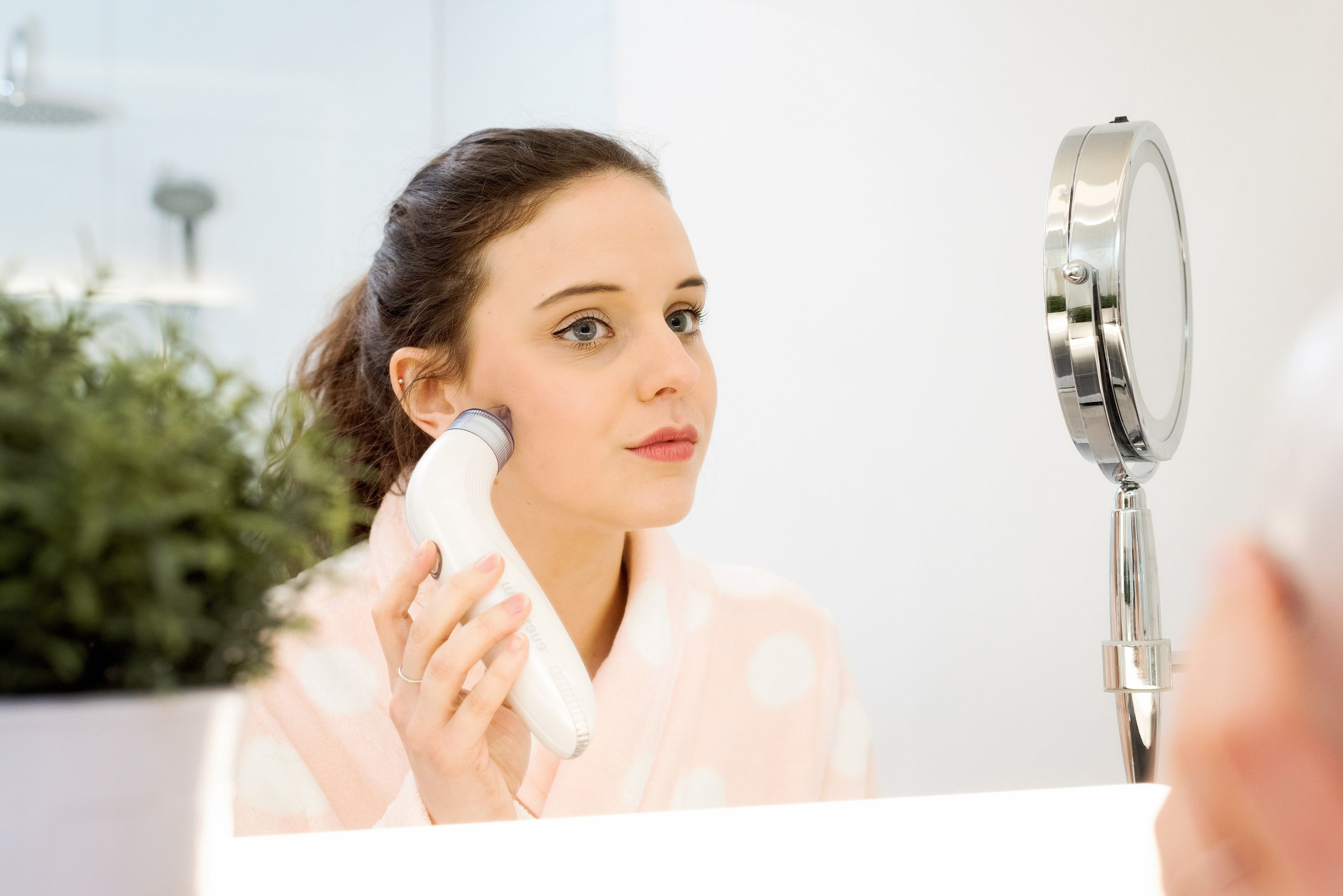 Mujer en bata usa un dispositivo de limpieza facial frente a un espejo.