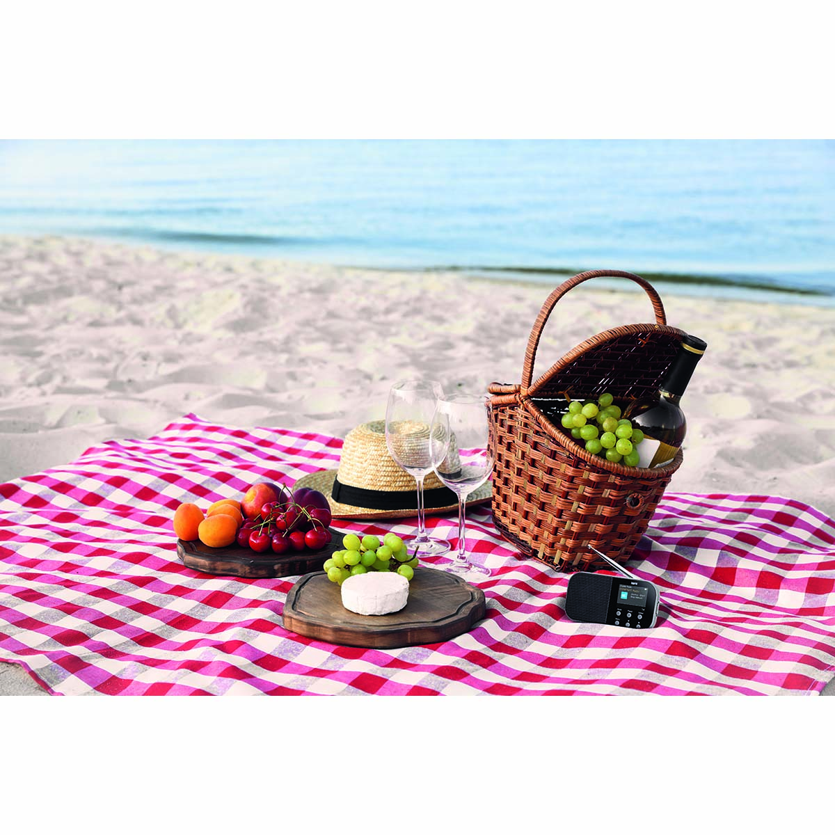 Scena piknikowa na plaży z jedzeniem, napojami i radiem na czerwono-białym pledzie w kratę.