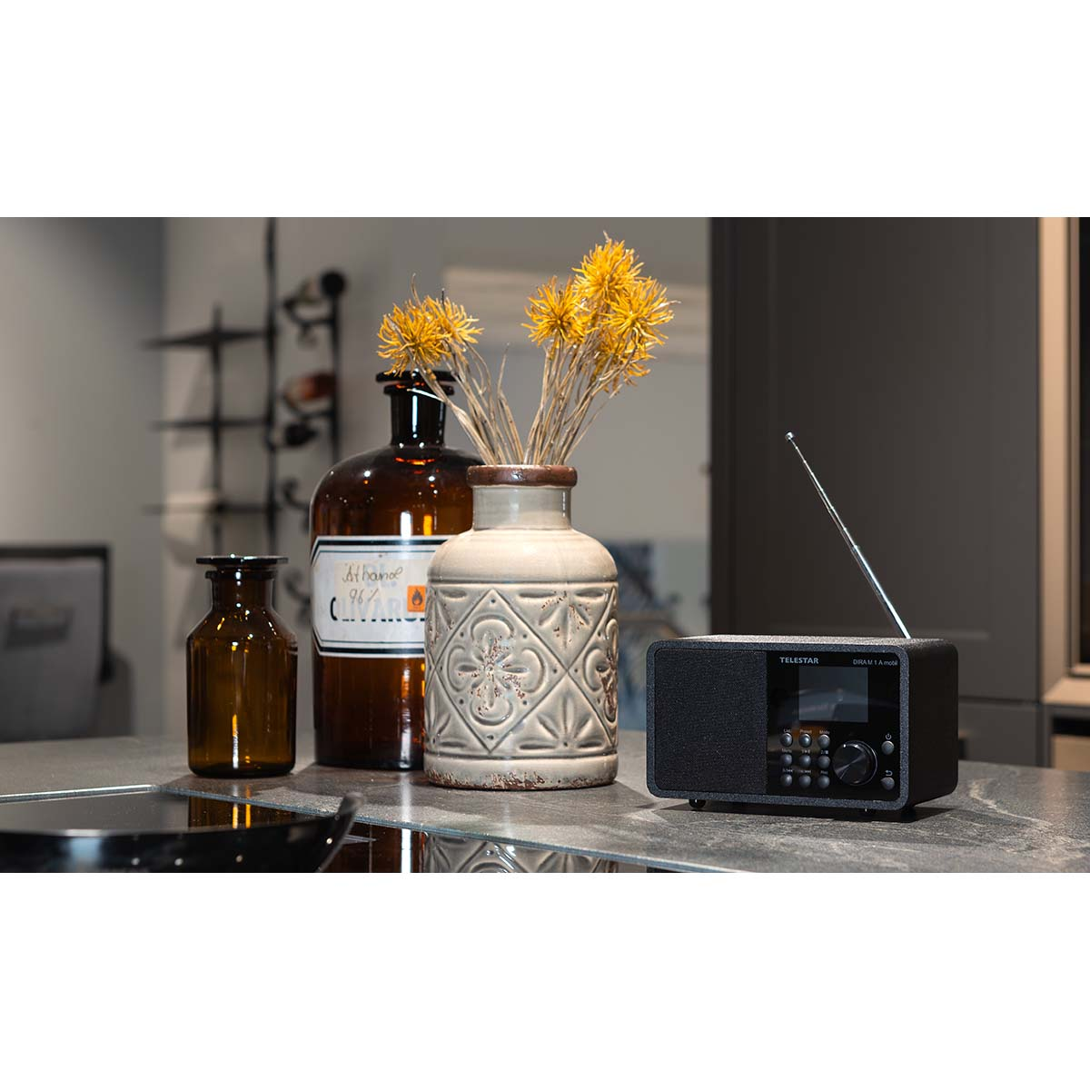A radio with a vase of flowers and two bottles on a counter. A gray wall is behind.