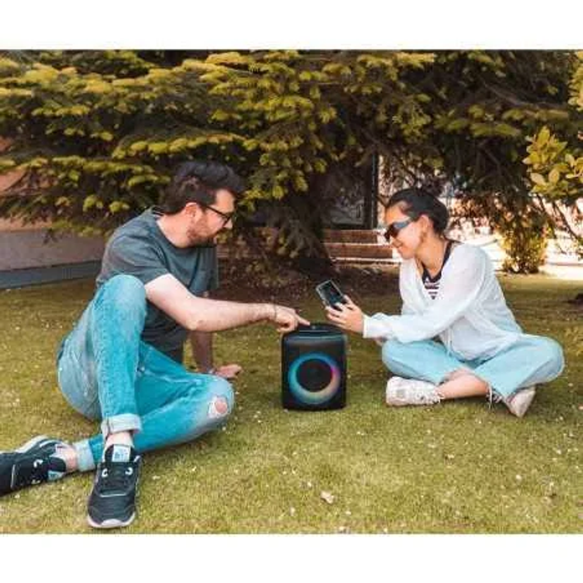Dos personas sentadas en la hierba, una señalando un altavoz con luz colorida, cerca de un árbol.