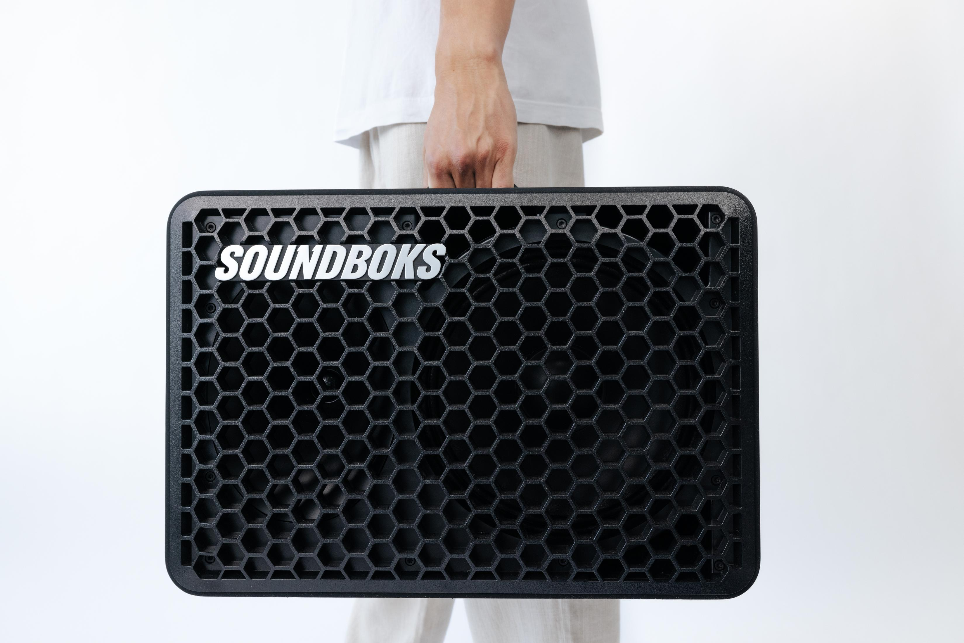 Eine Person hält einen großen, schwarzen SOUNDBOKS-Lautsprecher mit Wabenfront und SOUNDBOKS-Logo.