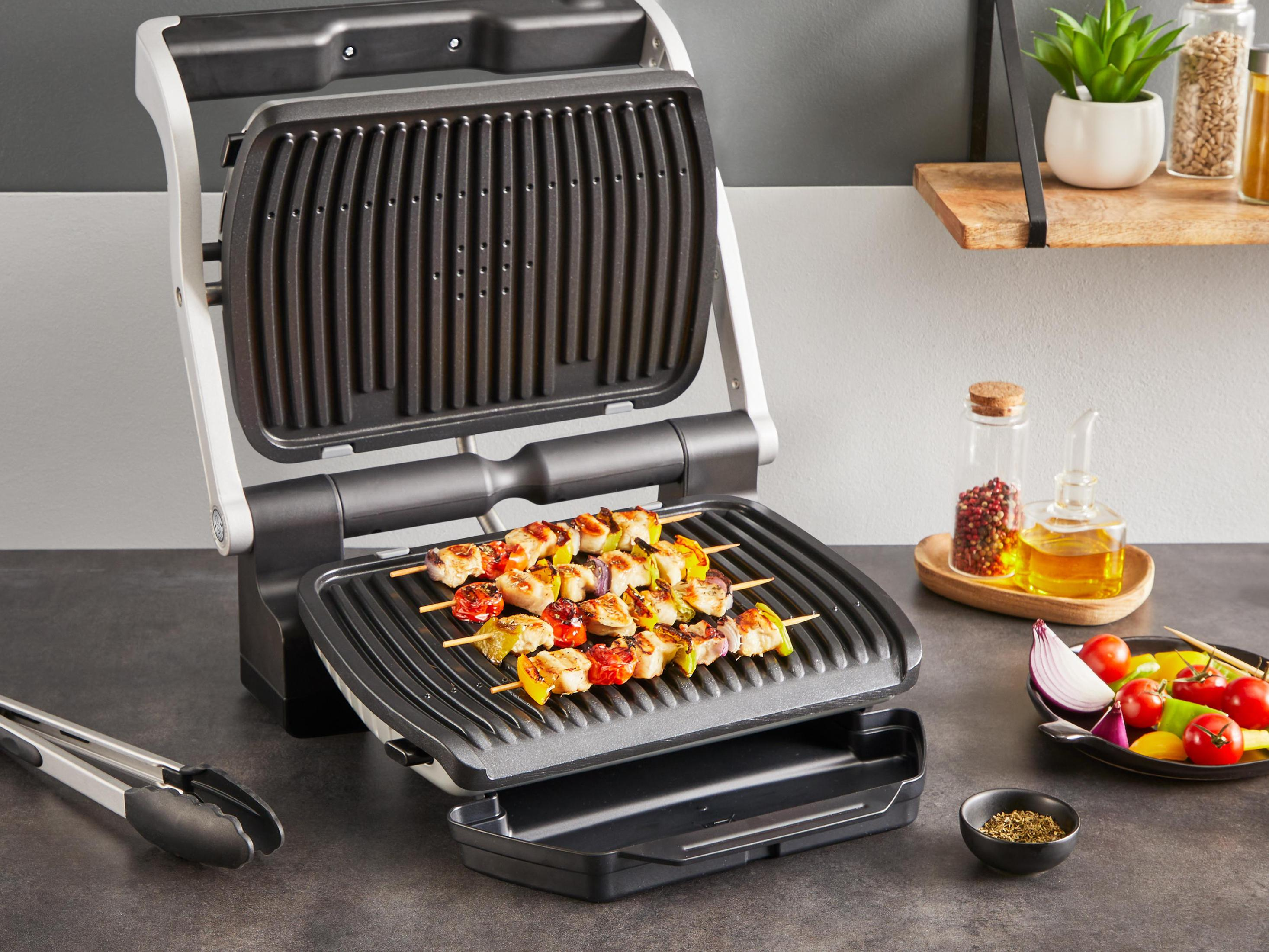 Offener Grill mit Essensspießen. Zangen und Zutaten stehen auf dem Tisch.