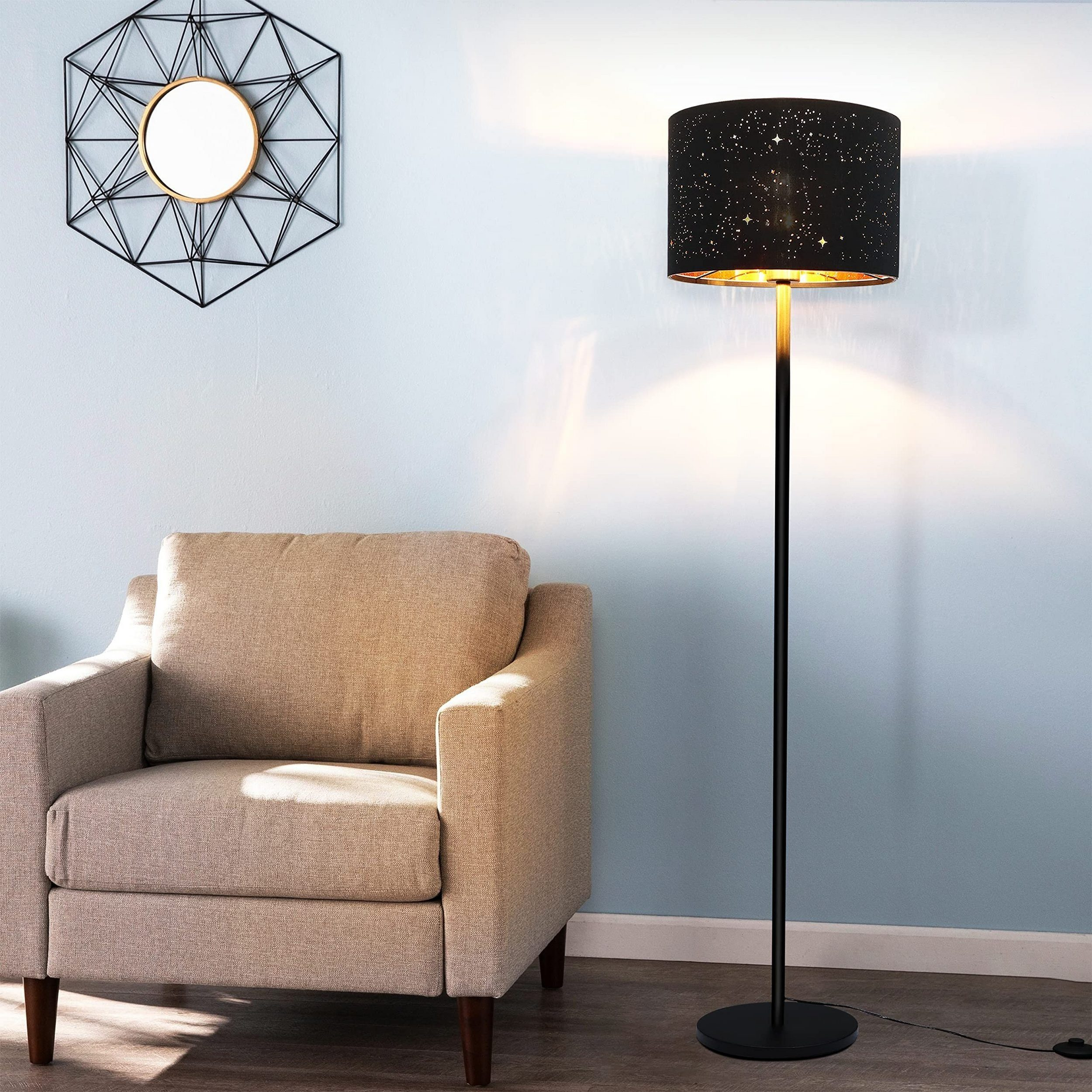 Czarna lampa podłogowa z gwiaździstym kloszem stoi obok beżowego fotela. Geometryczne lustro wisi na ścianie.