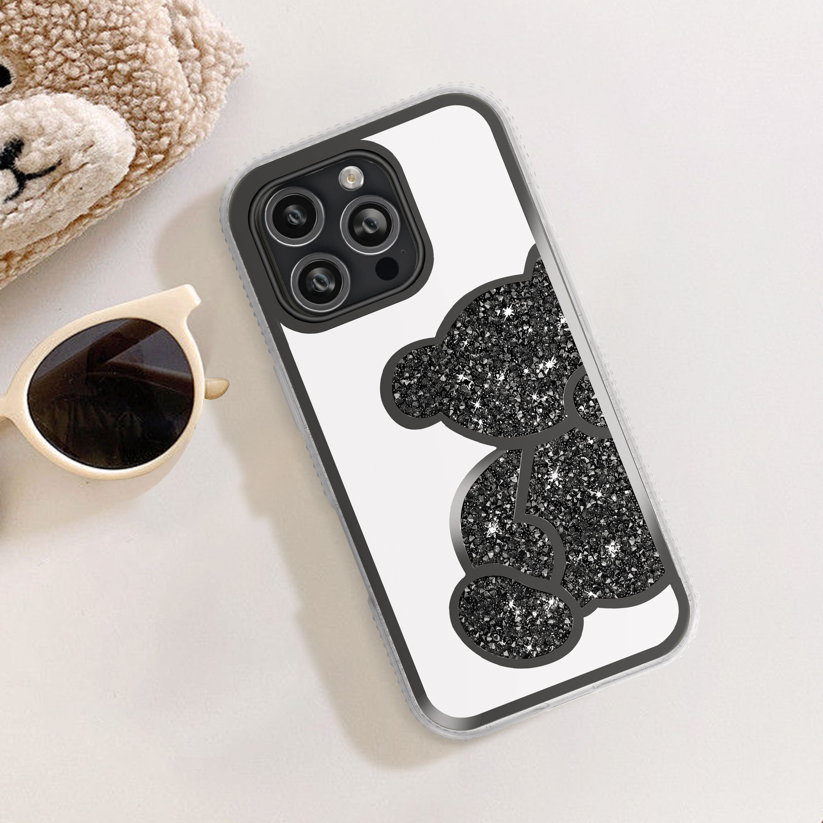 Una custodia per telefono con un design a orso nero scintillante, accanto a occhiali da sole e un orsacchiotto.